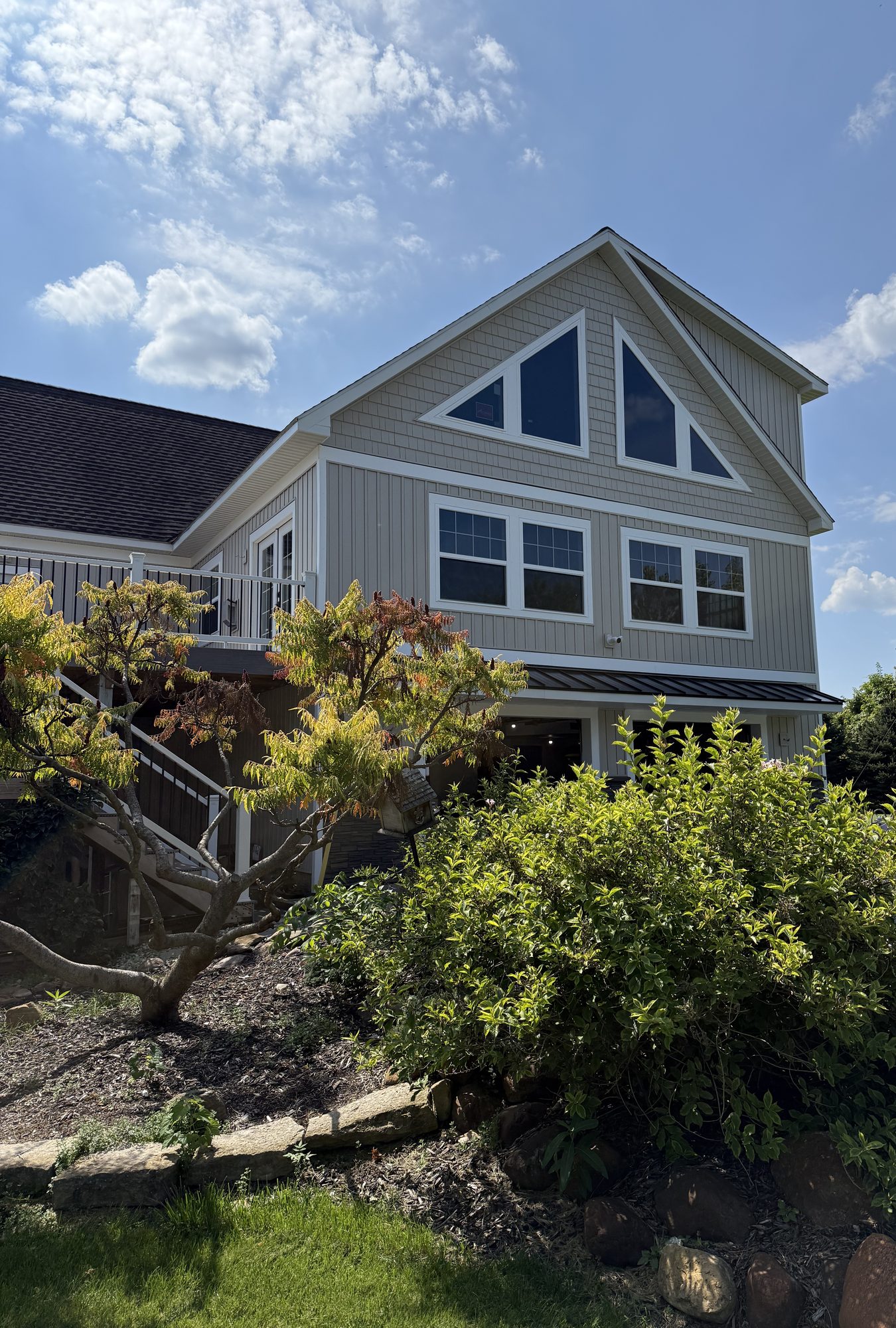Two-story home with new gray board-and-batten siding, large gable windows, and landscaped beds under a clear sky